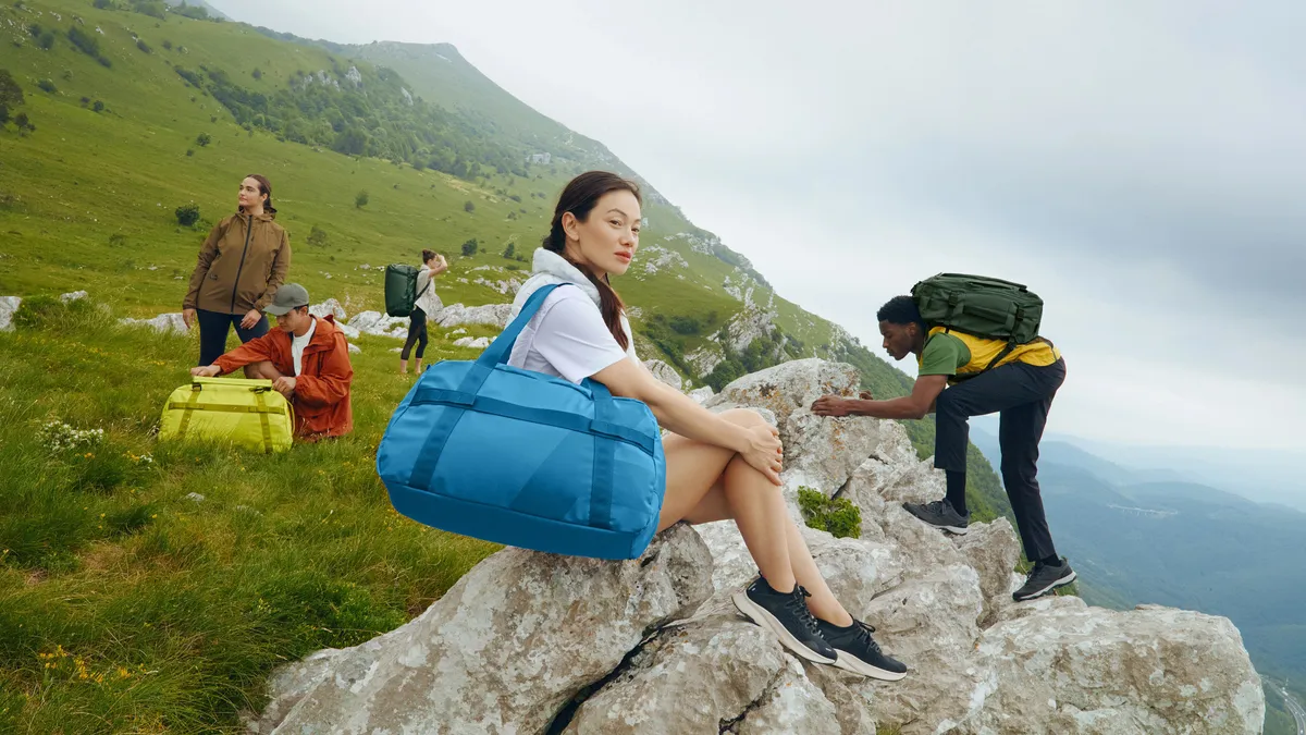 A diverse group of hikers deals with brightly colored luggage among large rocks on a grassy green mountainside.