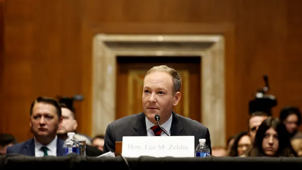 "Hon. Lee M. Zeldin," as a placard states, sits at a table with a microphone in front of a crowd in a hearing room.