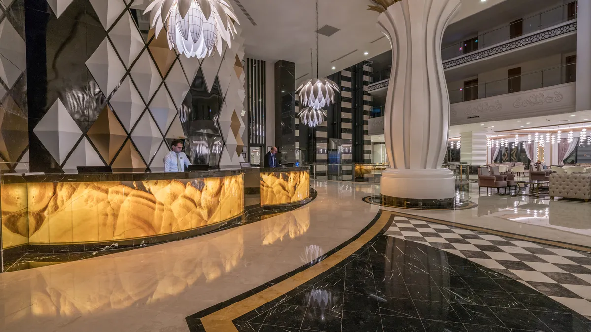 The lobby of a luxury hotel with two men behind an illuminated check-in desk and big white lighting fixtures overhead.