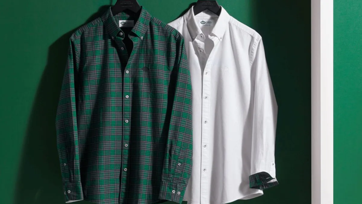 Two Oxford shirts, one in green plaid and the other white, hang on a Jets team color green background.