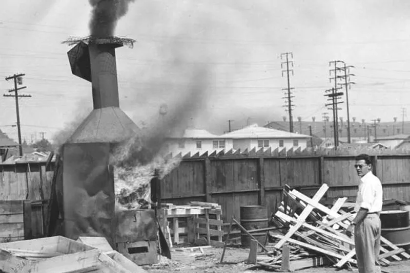 A man stands next to a small outdoor incinerator billowing smoke