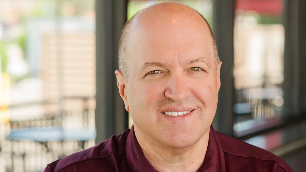 A smiling white man in a burgundy Noodles & Company shirt.
