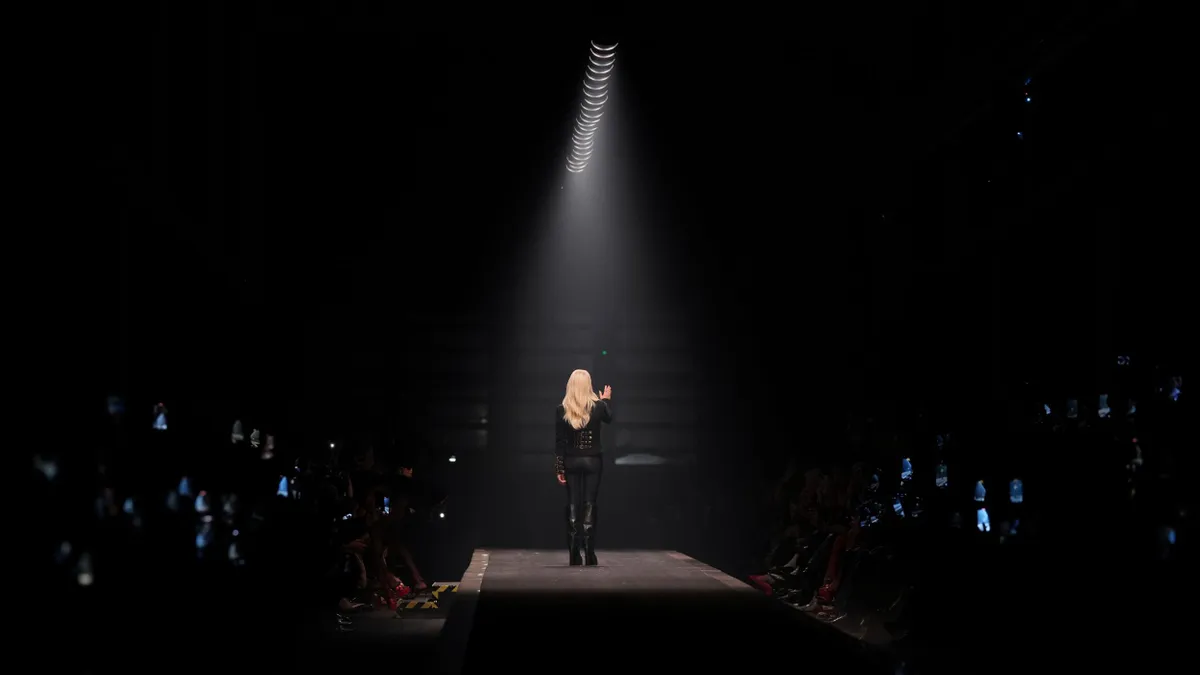 Donatella Versace waves under a spotlight on a runway.