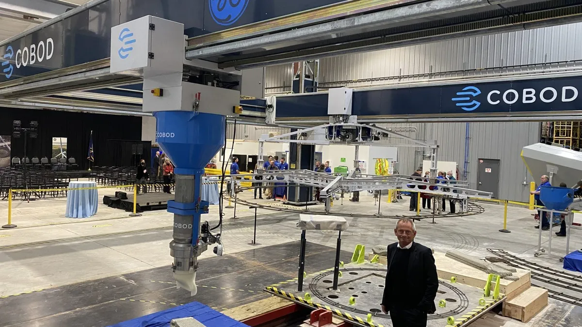 A man stands in the middle of a large, circular robot on rails in a warehouse.