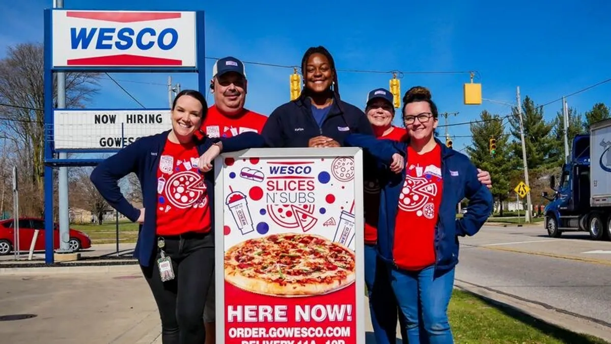 A photo of several people standing outside around a sign that reads "wesco slices 'n subs. Here now. Order.gowesco.com" while in the background a sign has the Wesco logo and a signboard below it reads "now hiring."