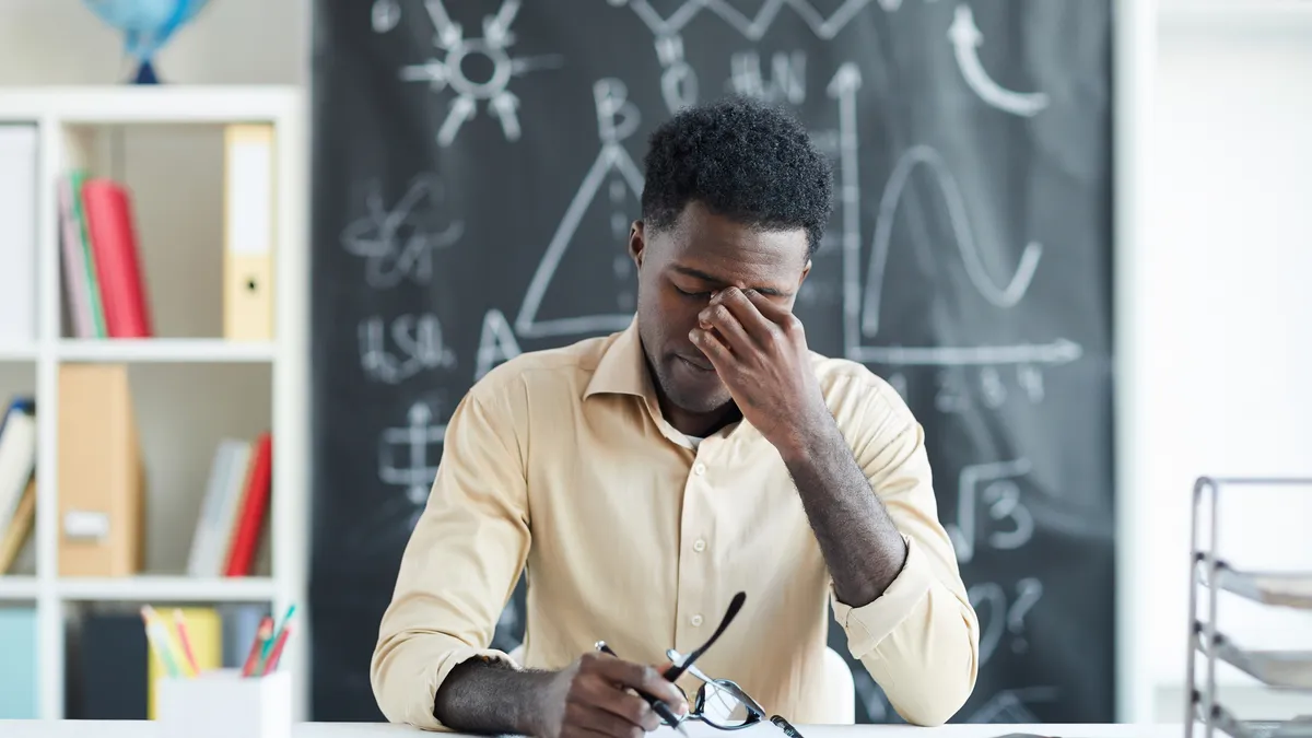 A teacher appears stressed out while sitting at a desk in their classroom. Behind the teacher is a chalkboard with writing on it.