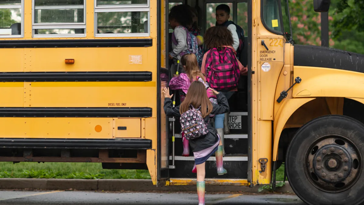 A yellow school bus is parked outside. A handful of students wearing rain jackets and  backpacks are getting on the bus.