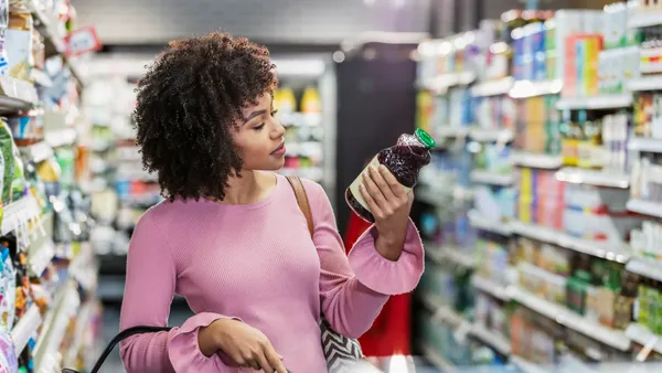 A person shopping in a grocery store, carrying a shopping basket and reading the ingredient label on a bottle.