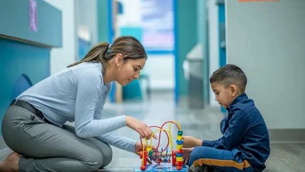 An adult is kneeling on the floor in front of a child. Both the adult and child are touching a toy with blocks and coils on the floor.