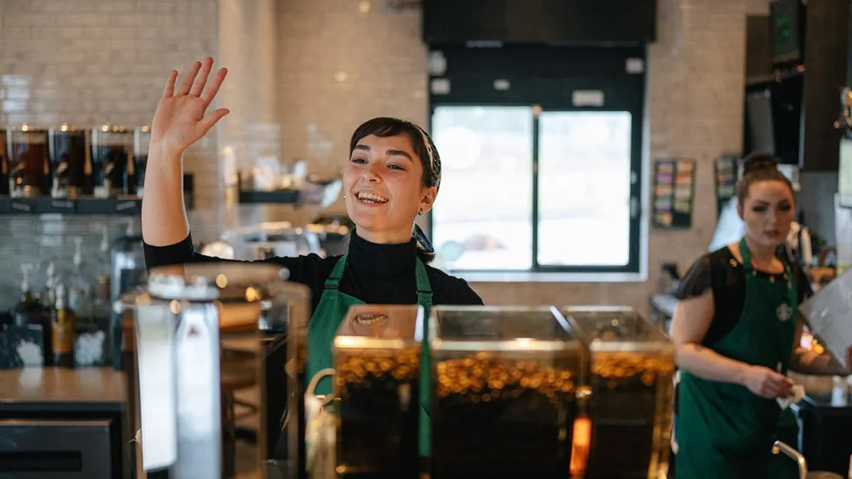 A woman in a green apron at a coffee counter waves off camera