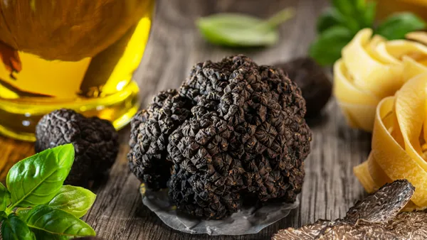 A truffle sits in the center of a wooden table surrounded by pasta, truffle shavings, basil and oil.