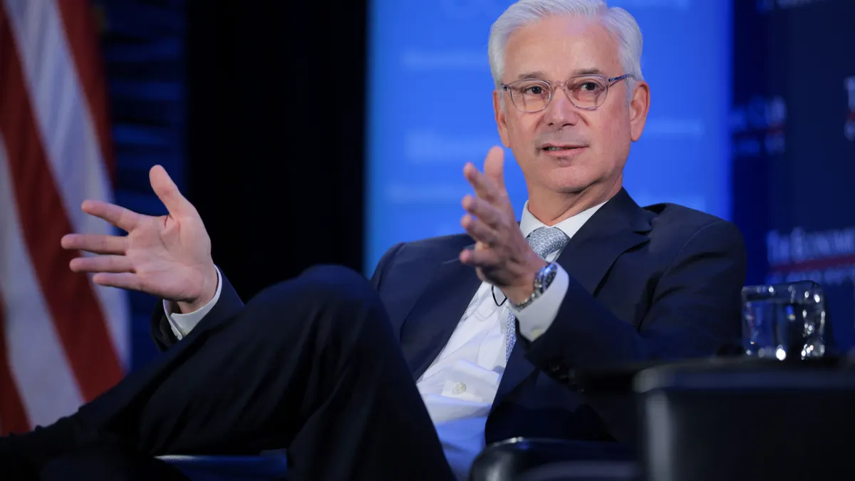 Wells Fargo CEO Charlie Scharf holds his hands out as he talks while sitting on a stage with a blue background. Part of an American flag is visible.