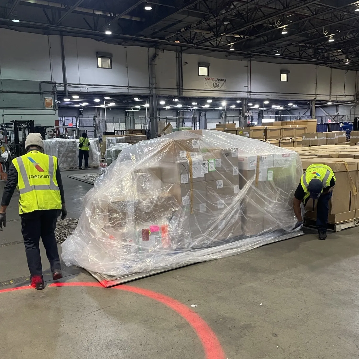 Workers in fluorescent yellow American Airlines vests wrap a pallet of products in plastic.