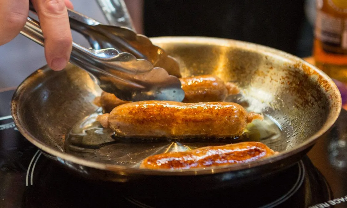 A person holds tongs to turn New Age Meats' cultivated and plant-based sausage as three links cook in a frying pan.