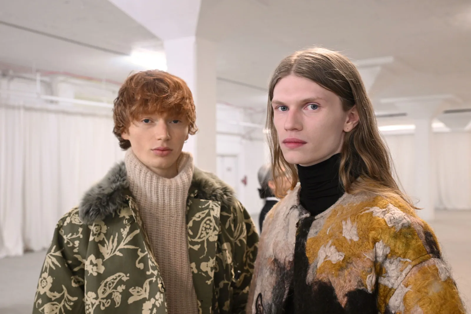 Two models wear patterned coats and stare at the camera while standing in a white lofty space.