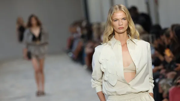 A model in a pale cream-colored blouse with a visible bra underneath walks on a runway.