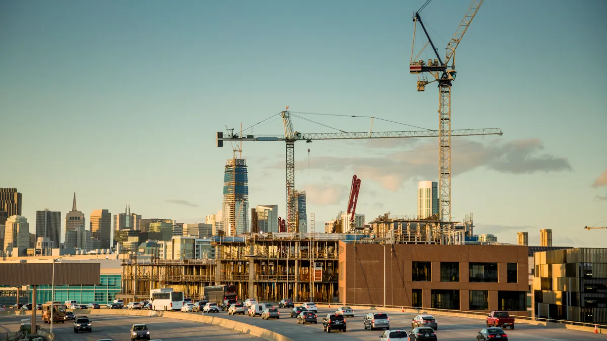 A highway with cars on it in front of a city skyline. Two large cranes line the highway next to new construction.