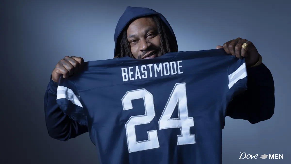 Former NFL player Marshawn Lynch holds up a football jersey with the name "Beastmode" inscribed across the back