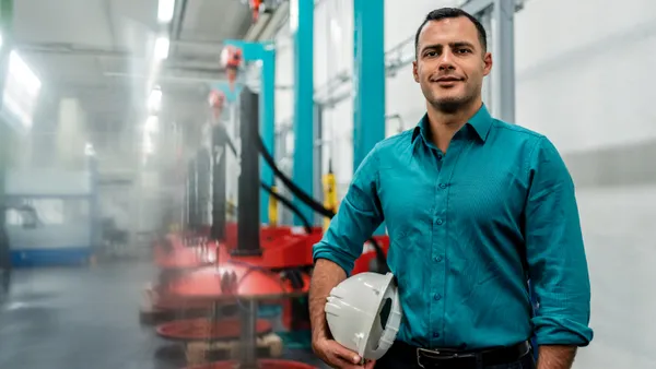 Smiling engineer with hands in pockets holding hardhat while standing in factory