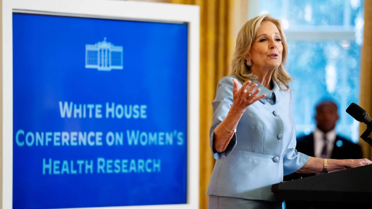 A person in a blue suit speaks at a podium in front of a sign reading "White House Conference on Women's Health Research."