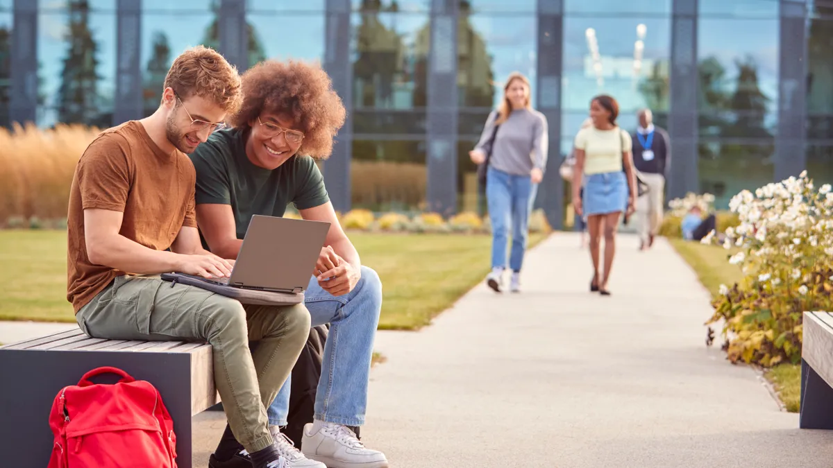 Male University Or College Students Sitting Outdoors On Campus Talking And Working On Laptop