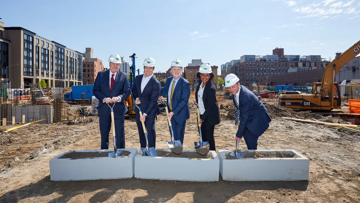 A group of people in formal business attire and hard hats turn dirt on a construction jobsite