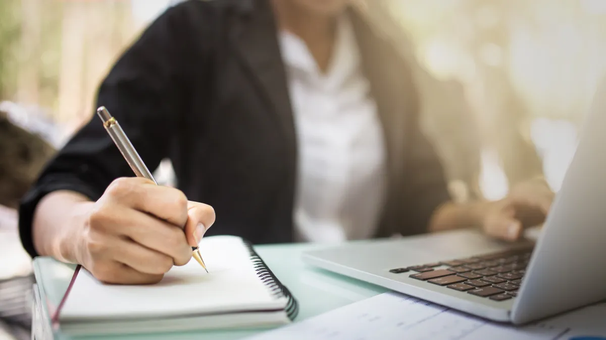 A person's mid section is shown sitting in front of a laptop. A hand is holding a pen and writing in a notebook.