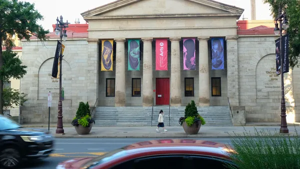 View of building with columns and colored banners as vehicles and a pedestrian pass.