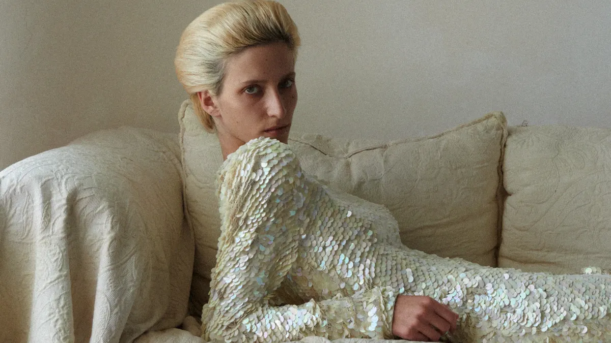 A person in a long-sleeved cream sequined dress reclines on their elbows and stares at the camera.