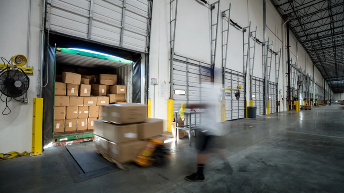 A busy warehouse interior with open loading dock doors. A worker moves cardboard boxes on a pallet jack, creating a sense of movement and efficiency.