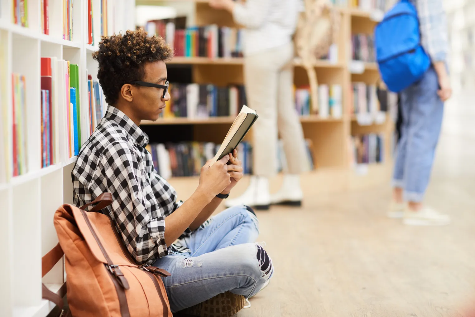 A serious teenager sits with crossed legs on a library floor, reading an interesting book.