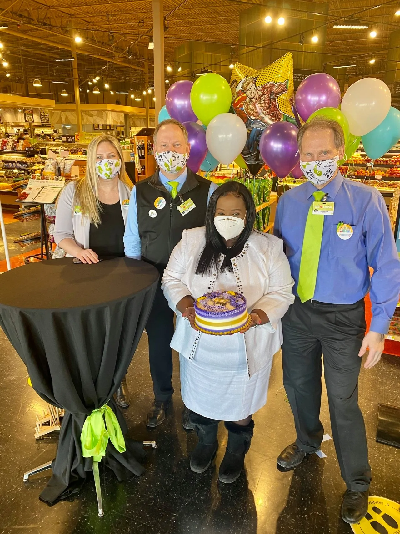 Four people wearing face masks in a grocery store with purple, green, blue and white balloons behind them. One person is holding a cake.