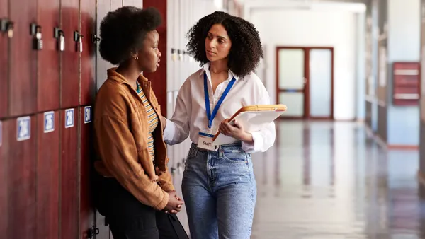 A student and teacher in a school hallway with lockers are talking