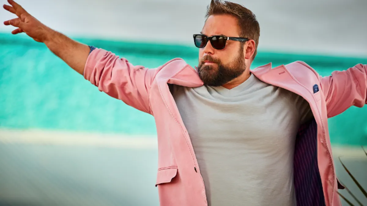 Person wearing a pink blazer and gray shirt and sunglasses with arms outspread.