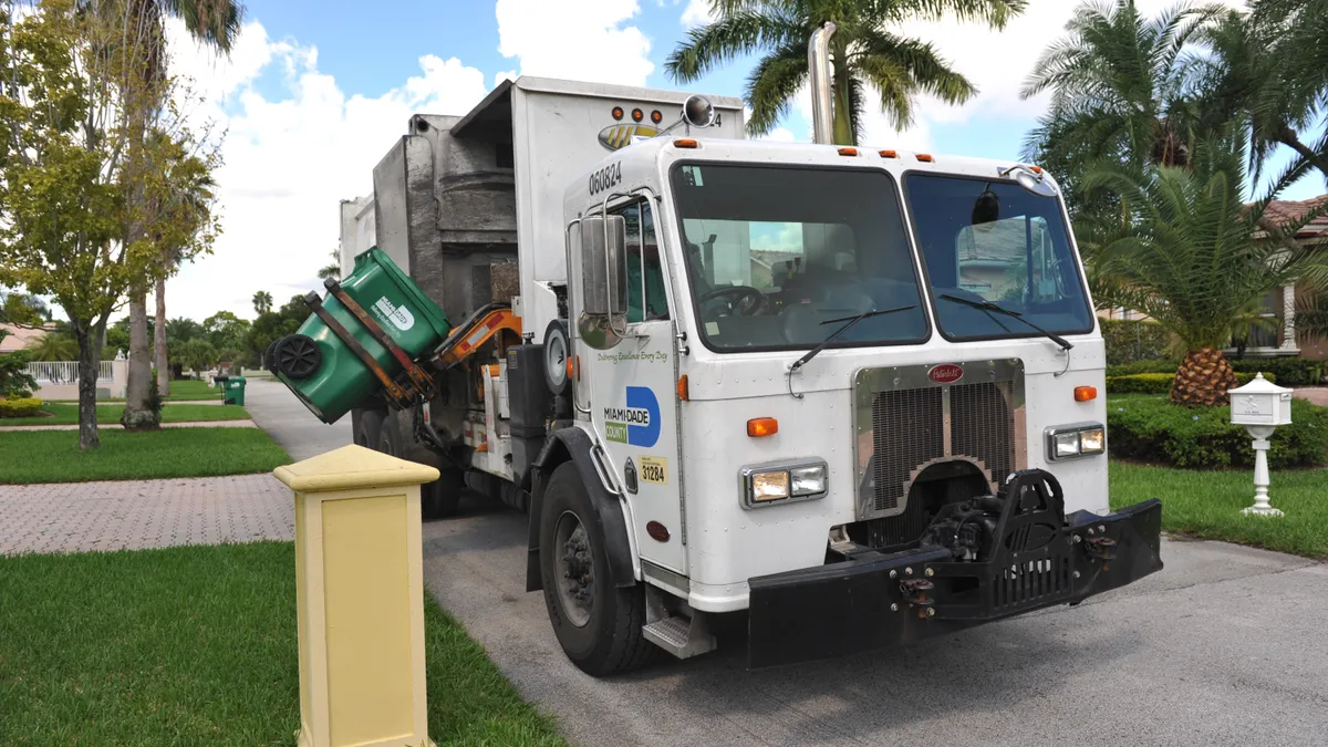 A Miami-Dade County side-loading waste collection truck picks up a garbage bin on a street.