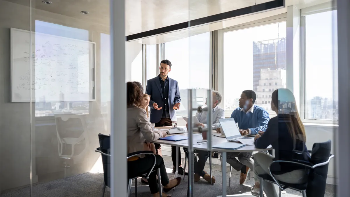 an executive in business attire leads a meeting of five workers who are seated in a bright conference room.