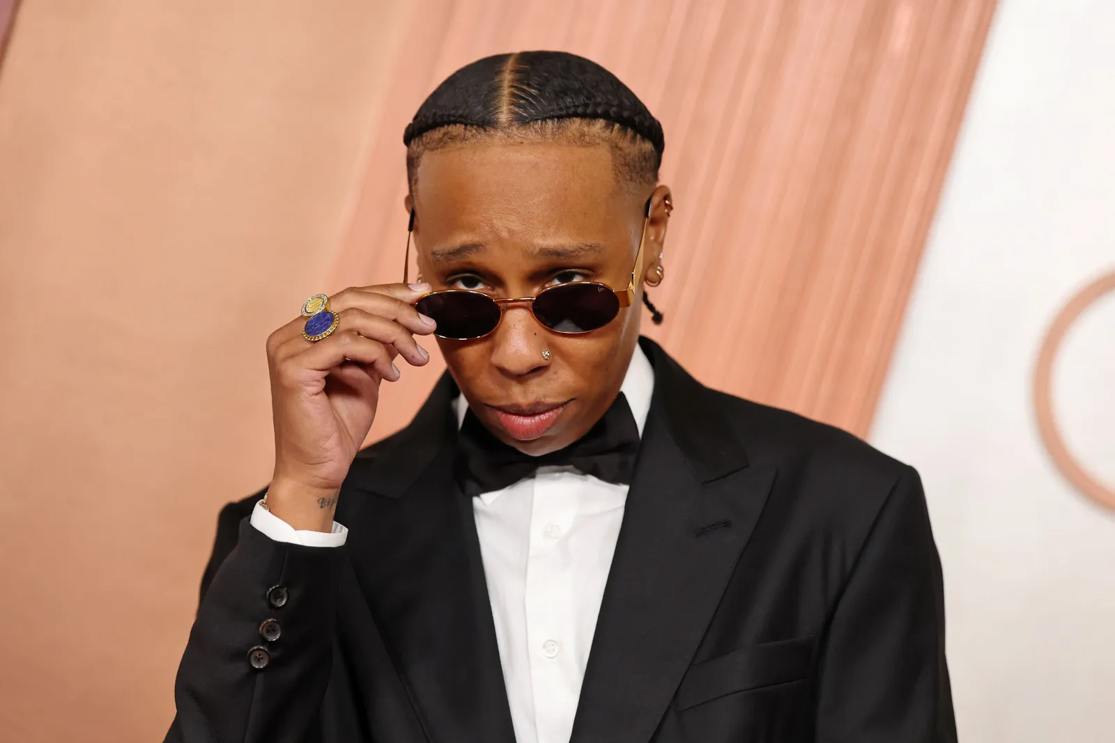 A woman in a classic tuxedo peeks at the camera over a pair of small round sunglasses.