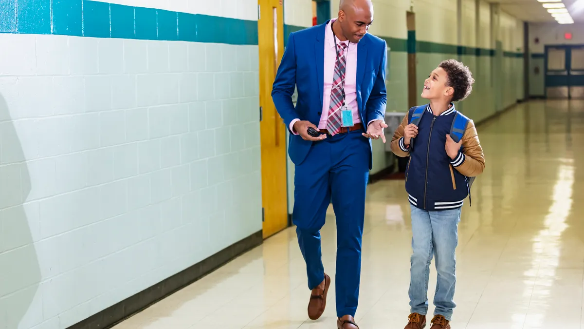 A principal dressed in a suit walks down a school hallway with an elementary student walking on the left side of the administrator.