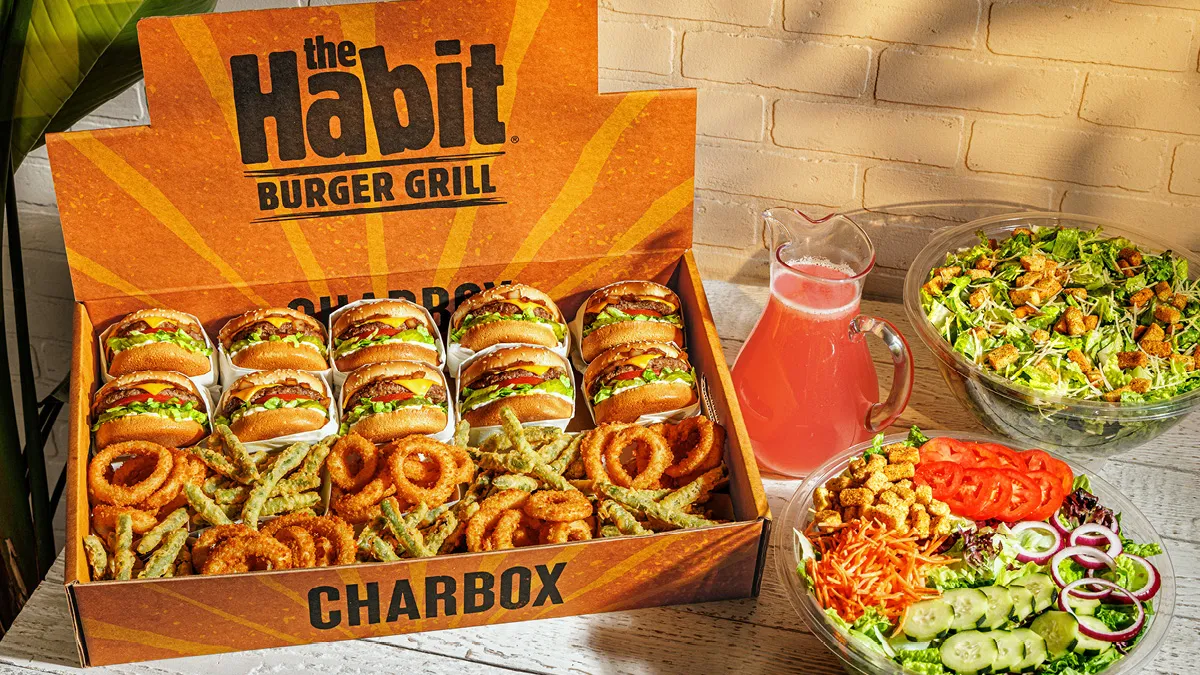 An image of a box with 10 burgers and sides next to a pitcher of pink liquid and a large salad. The box has "The Habit Burger" on it.