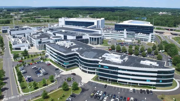 A campus with white and glass buildings surrounced by a parking lot and trees.
