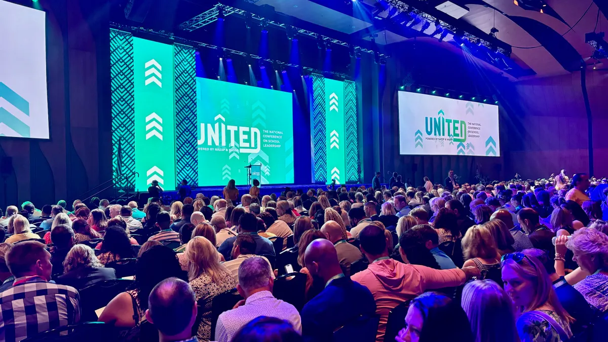 A large crowd of people seated are facing a stage. On the wall behind the stage are several large lighted signs that read UNITED.