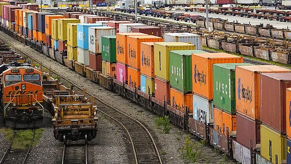 Container freight is being transferred between rail and truck at an intermodal facility where several colored containers can be seen.