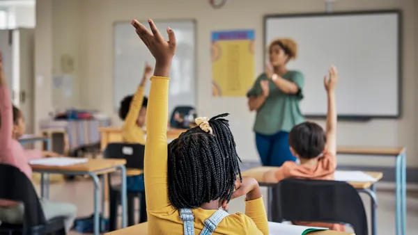 Students sitting in desks in a classroom have their backs to the camera and their arms raised. An adult stands in the front of the classroom facing the students.