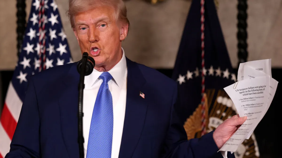 President Donald Trump holds up a few papers while speaking into a microphone.