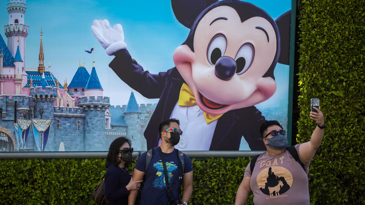 Three people in masks take a selfie on an iPhone in front of a Disneyland Park poster.