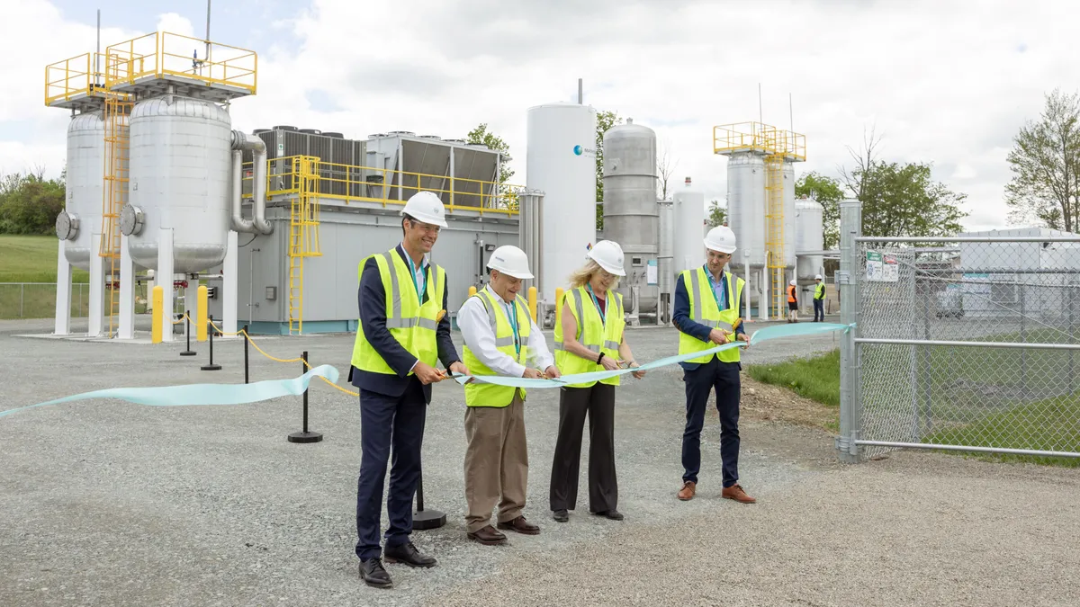 Four people wearing construction vests and hats cut the ribbon in front of a small Waga Energy industrial facility with tanks and pipes on a breezy day.