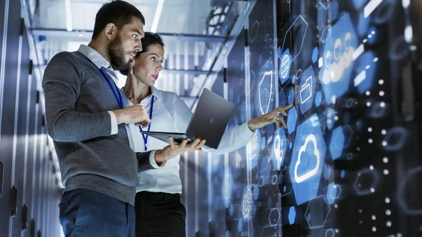 A male IT specialist holds a laptop and discusses work with a female server technician in a data center, standing before a rack server cabinet with a cloud server icon and visualization.