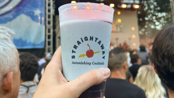 A person's hand holds a plastic cup with branding for Straightaway Cocktails, and an outdoor concert stage is in the background.