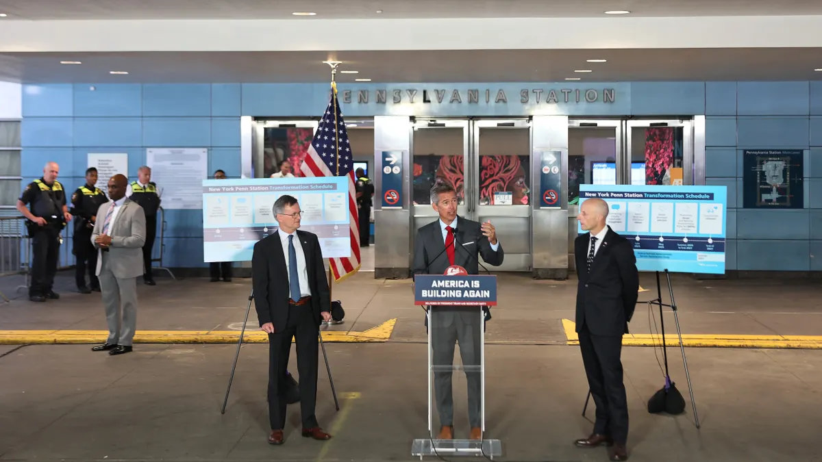 Three men in suits, one standing at a podium, with an American flag and a sign reading Pennsylvania Station behind them.
