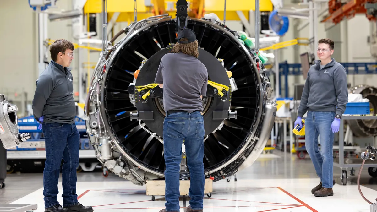 Three people in gray sweaters, blue jeans and protect eyewear working on a plane engine.